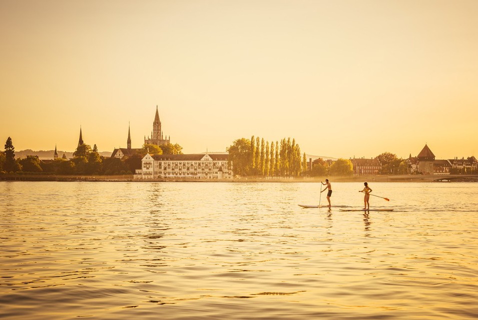 Stand-Up Paddleboarding on Lake Constance at Sunset Two persons stand-up paddleboarding on the calm waters of Lake Constance, surrounded by the warm glow of sunset