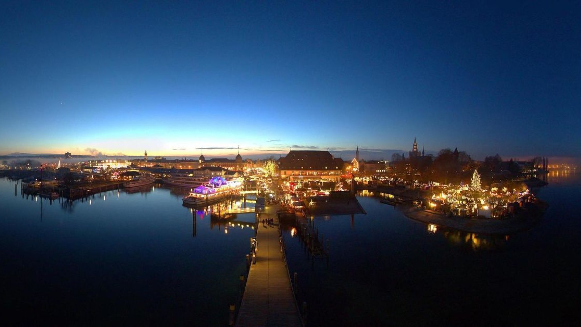 The brightly lit Christmas market in Konstanz at night, seen from the lake, with festive lights reflecting on the water